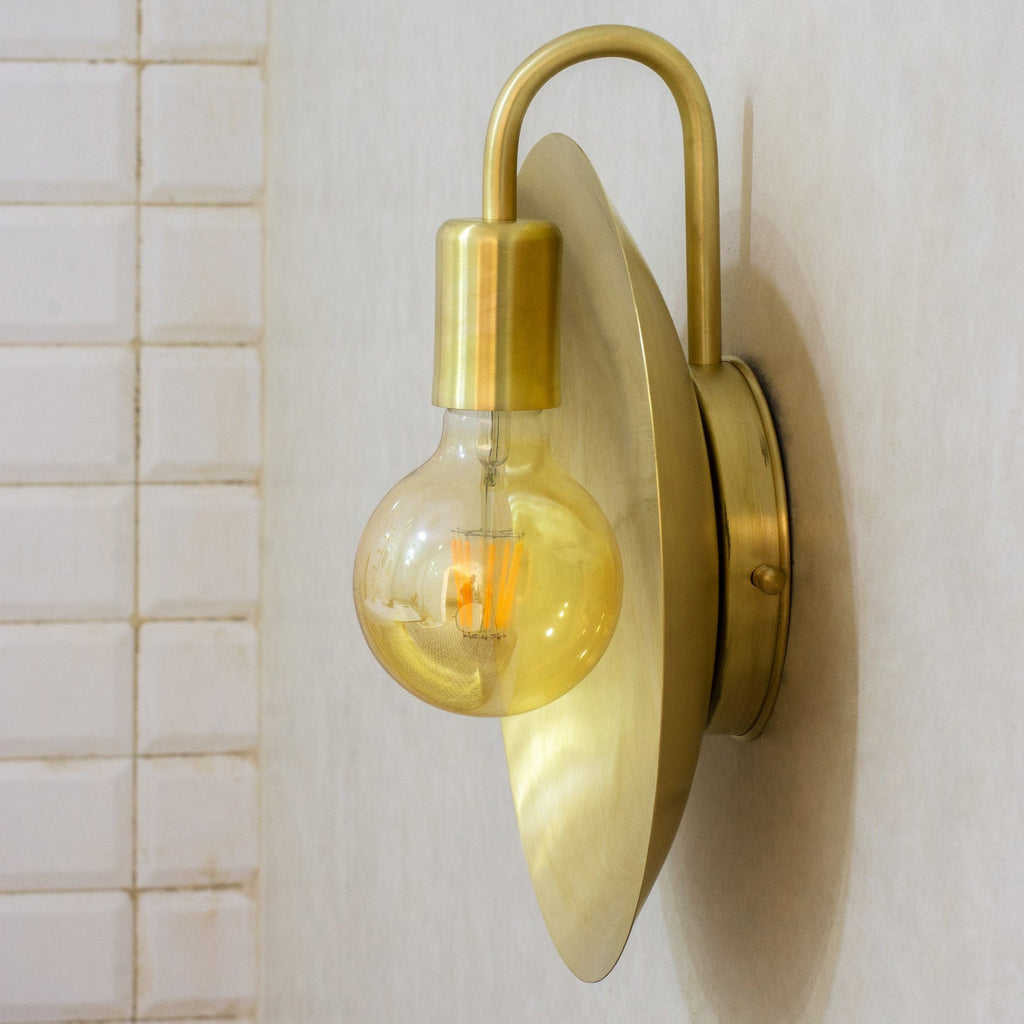 Angled view of a brushed brass dish wall sconce with a curved arm and exposed bulb