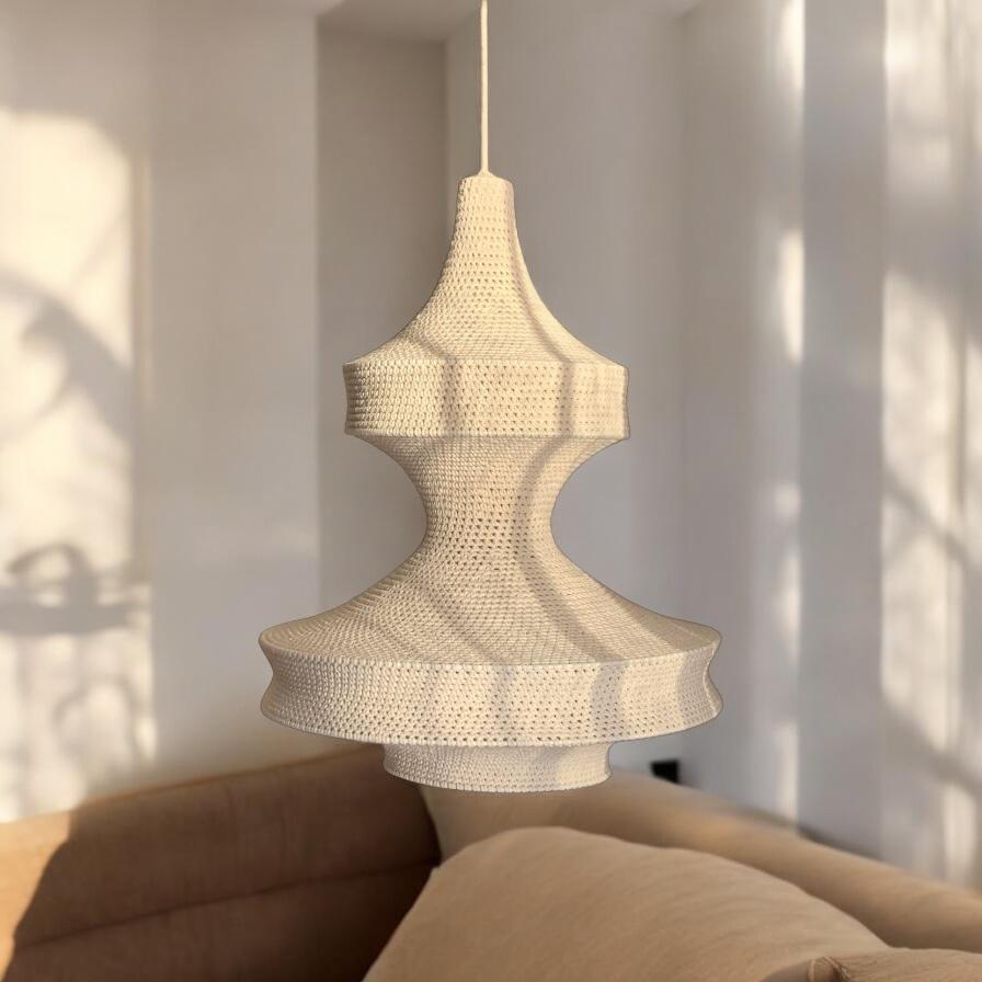A cream-colored pendant light fixture featuring a multi-tiered, bell-shaped shade constructed from a fine, intricate…