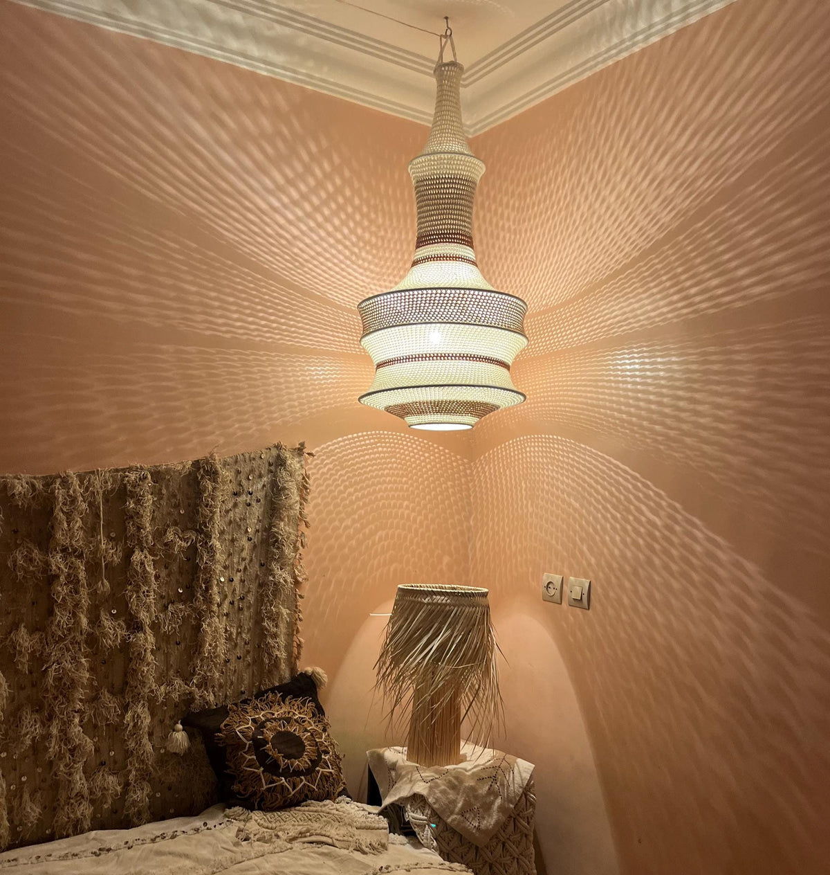 A tall, bell-shaped pendant light fixture with a woven, off-white texture hangs from the ceiling, casting intricate,…