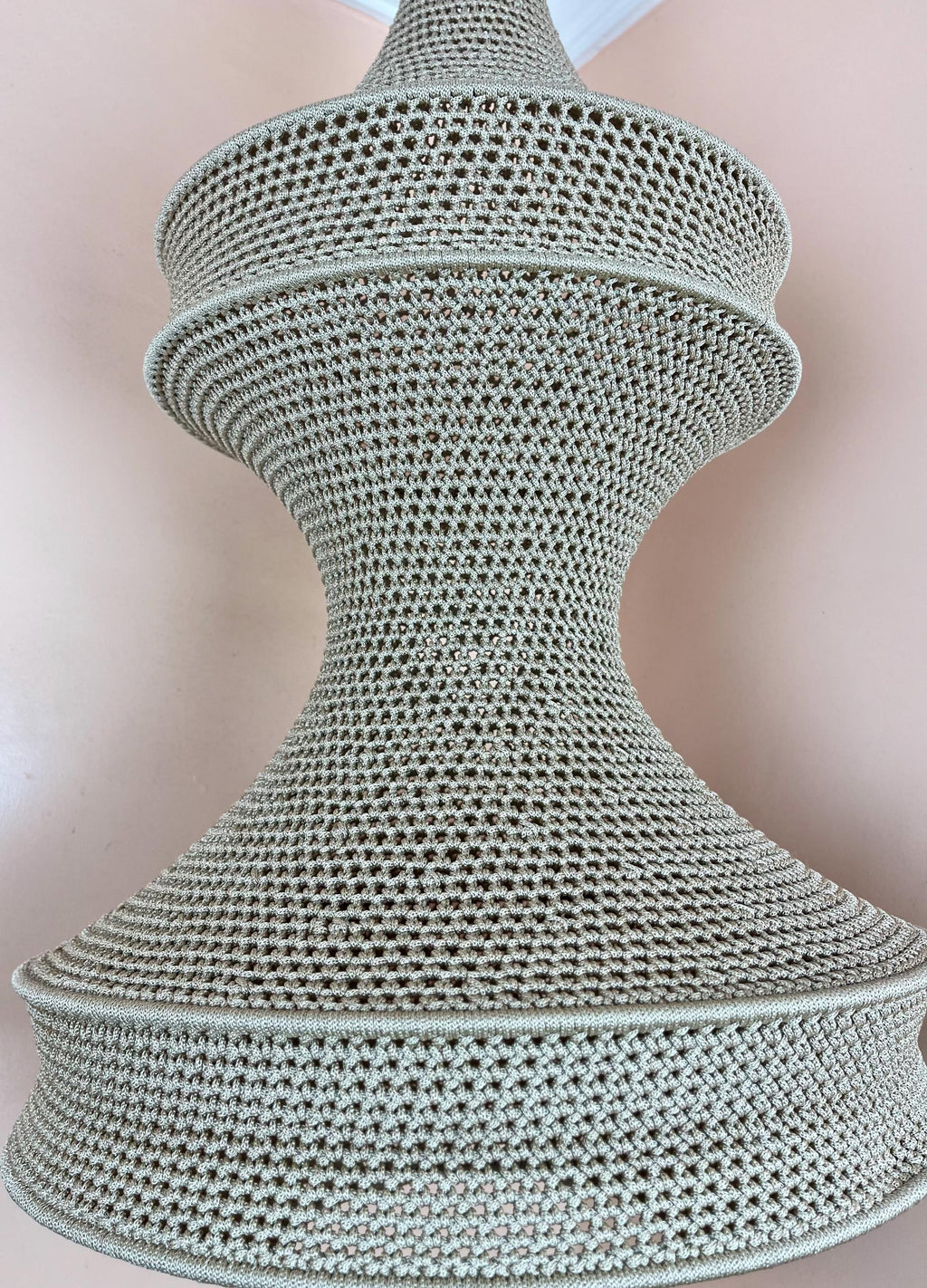A beige crochet lampshade with a distinct hourglass silhouette, featuring a fine, uniform mesh pattern throughout. The…