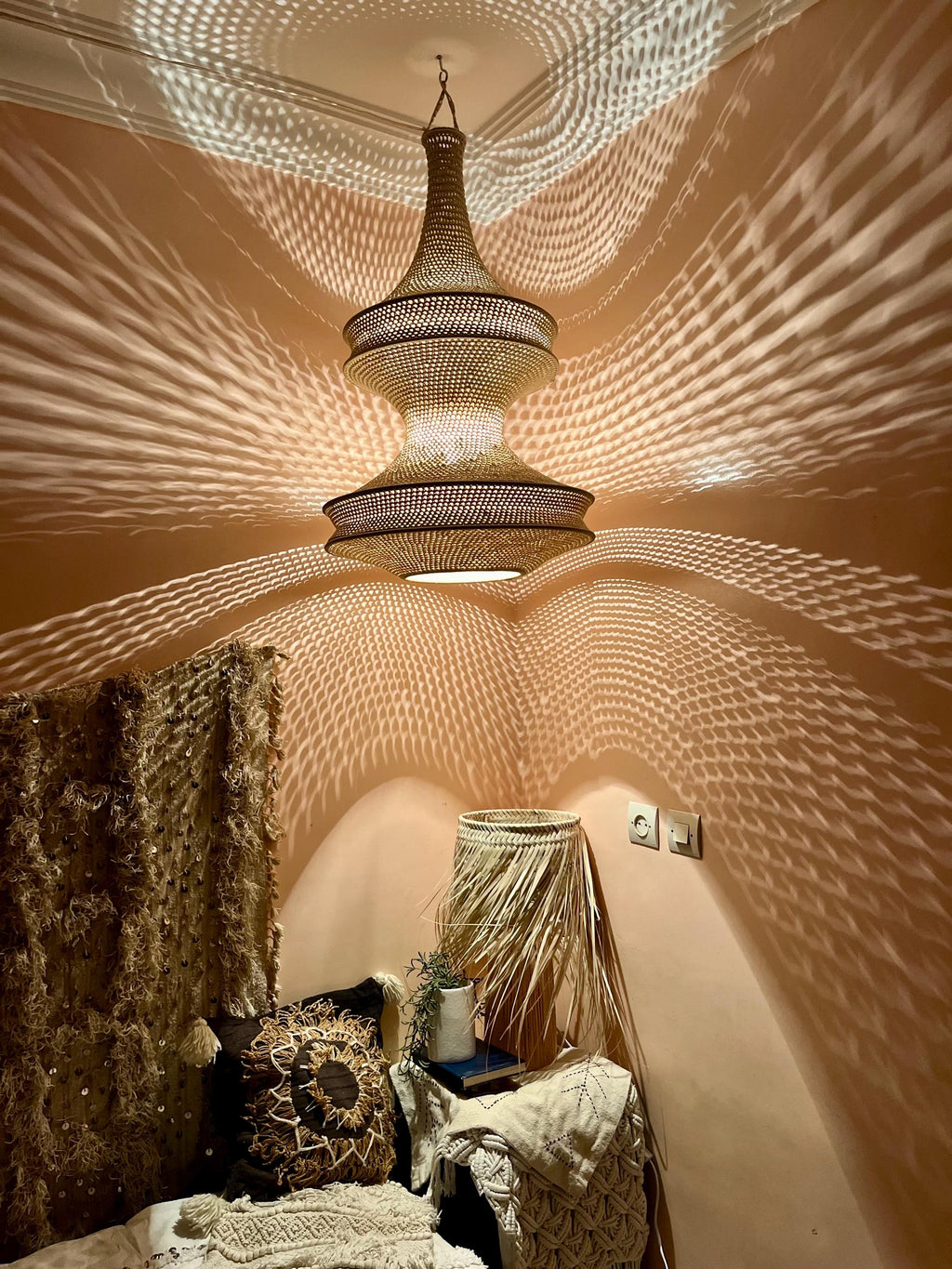A hanging pendant light fixture with a woven, tiered, hourglass-shaped shade made of natural fibers. The light emits a…