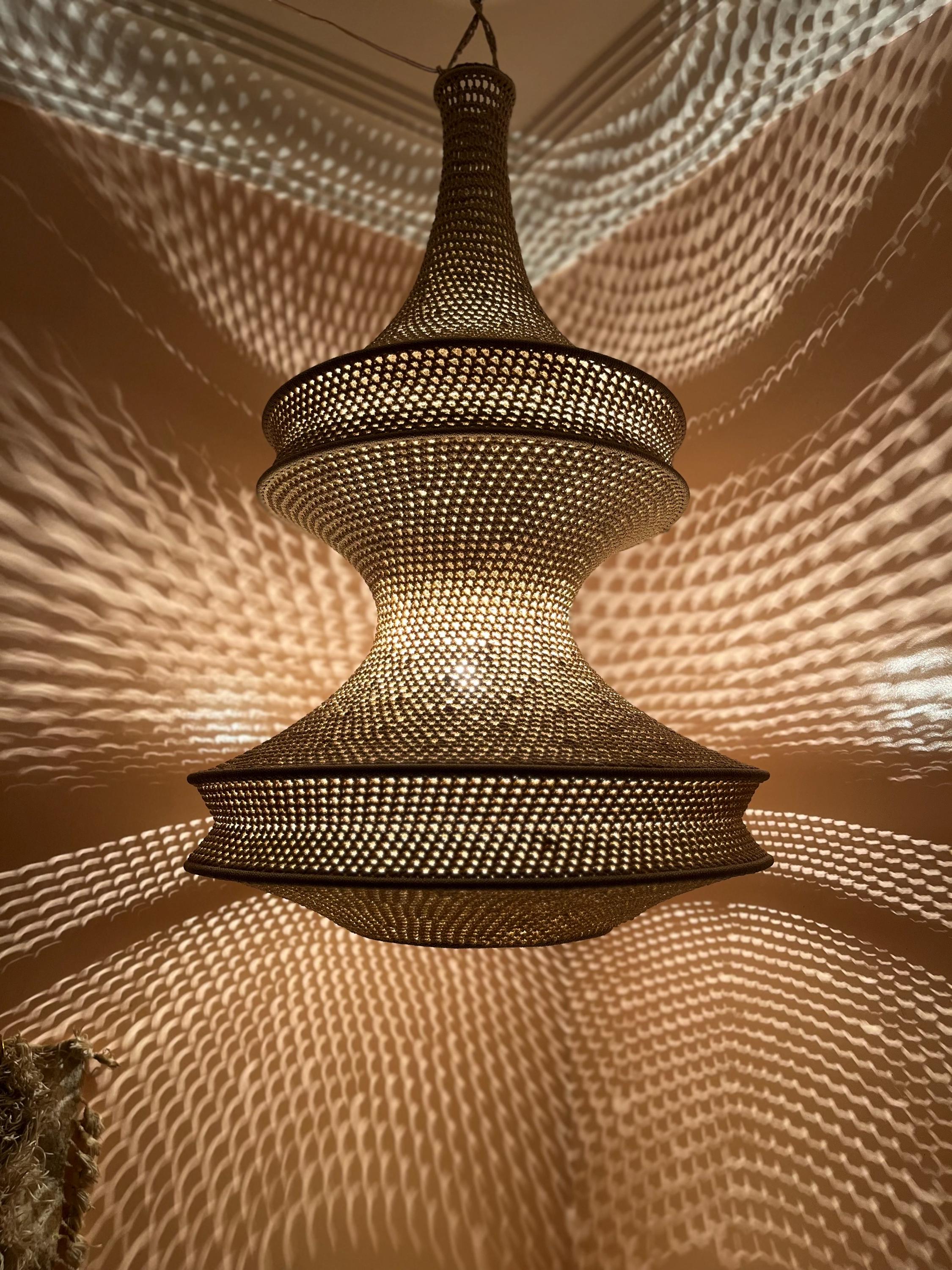 A beige woven pendant light fixture with a tiered, hourglass-shaped silhouette. The intricate mesh structure allows light…