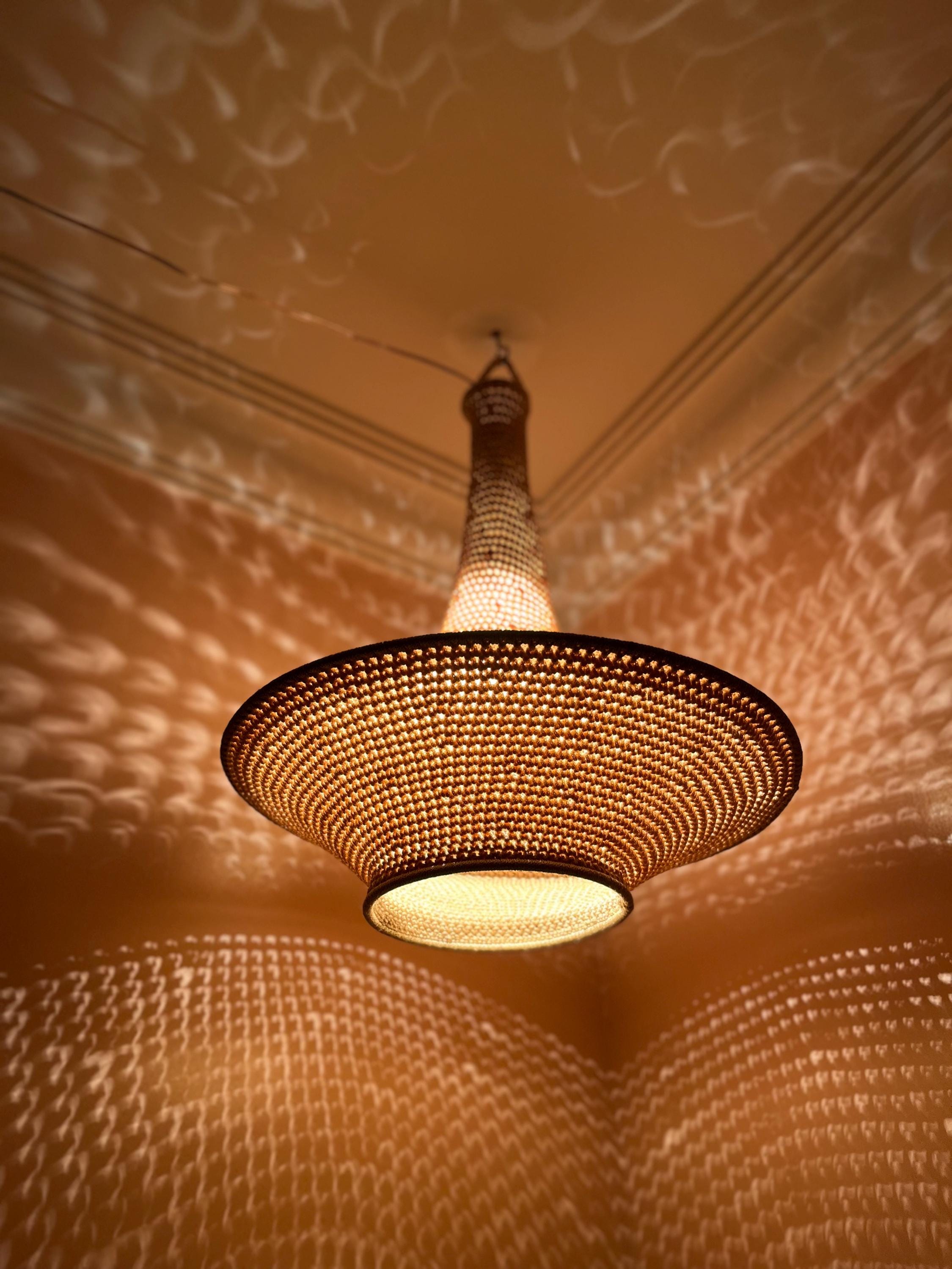 A bell-shaped pendant light fixture crafted from woven natural fibers in a warm tan hue. The structure features a narrow…