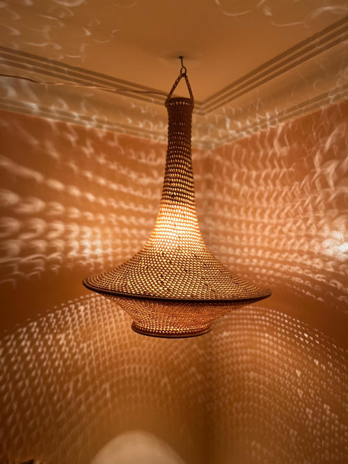 A hanging pendant light fixture featuring a woven, tan-colored material with a wide, flared base and a tall, narrow neck.…