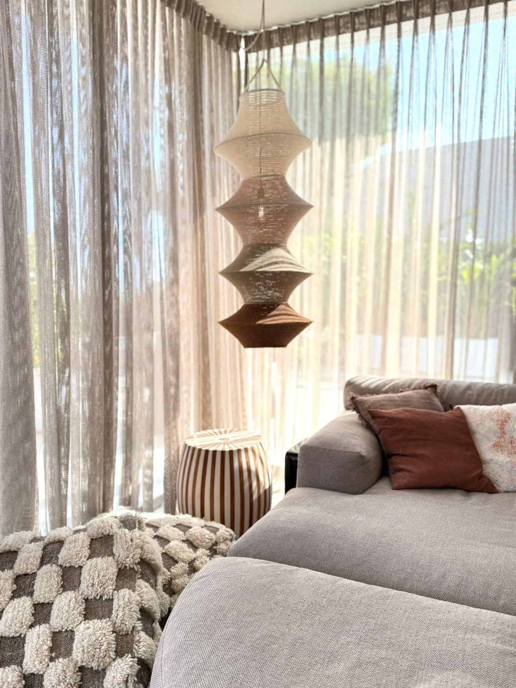 A hanging pendant lamp featuring four stacked, tapered fabric shades in a gradient of colors ranging from cream at the…