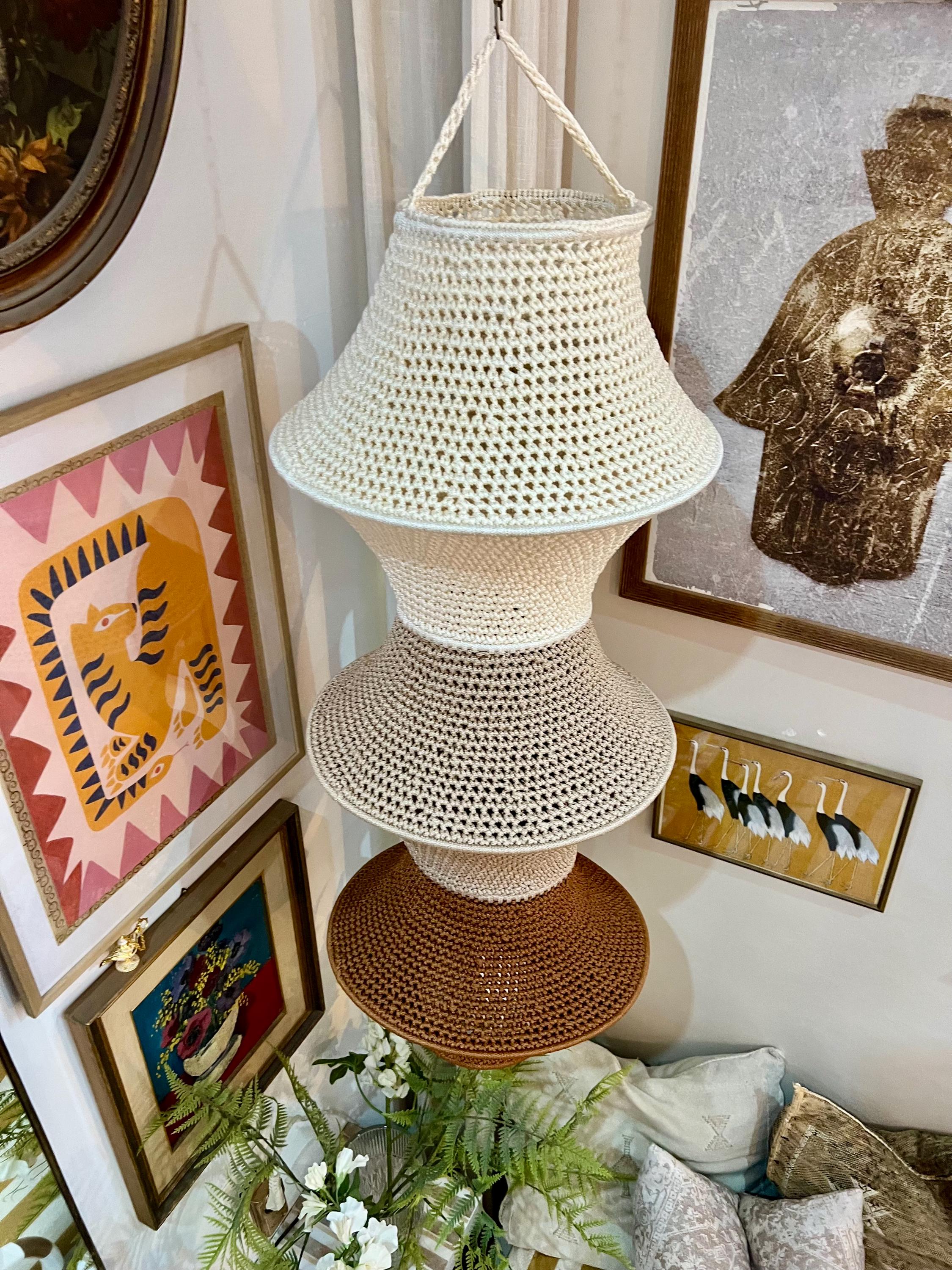 A hanging pendant light fixture featuring a tiered, crocheted shade design. The structure consists of three stacked,…