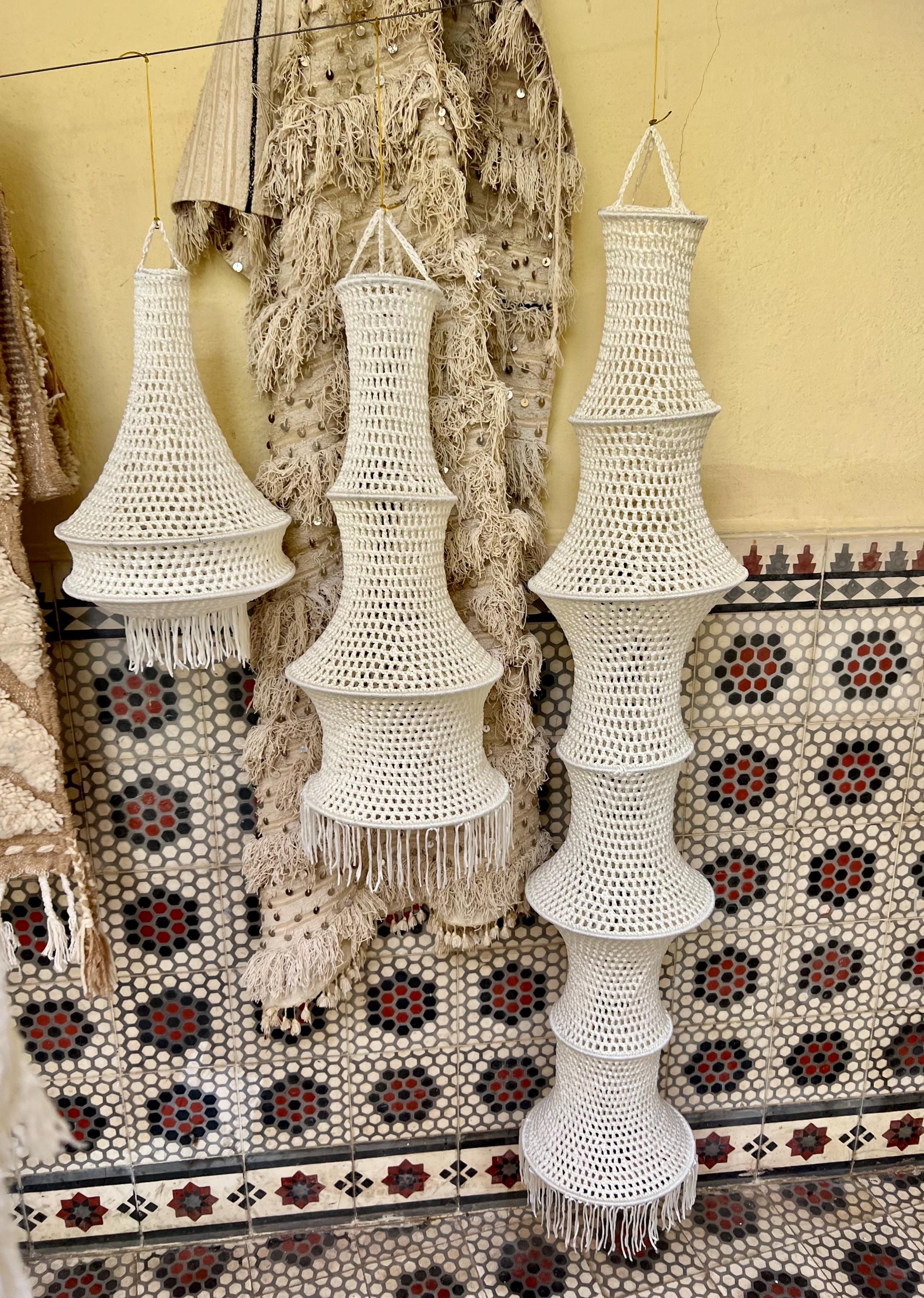 Three off-white crocheted hanging lampshades of varying heights and tiered shapes. Each shade features an open-weave…