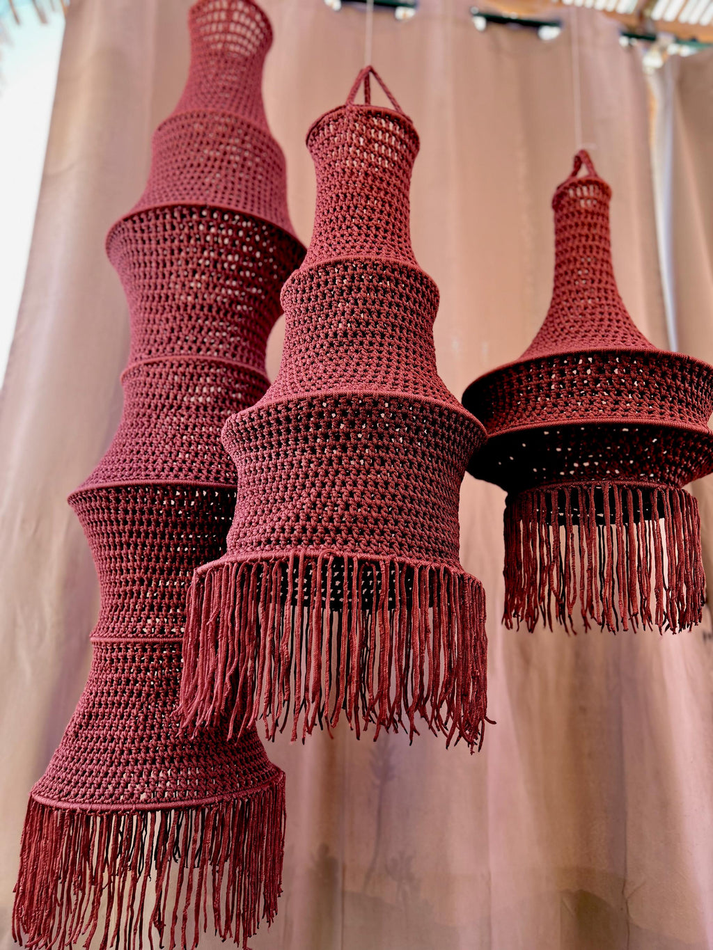 Three hanging pendant lamps crafted from dark red crocheted yarn. Each lamp features a tiered, bell-shaped structure with…