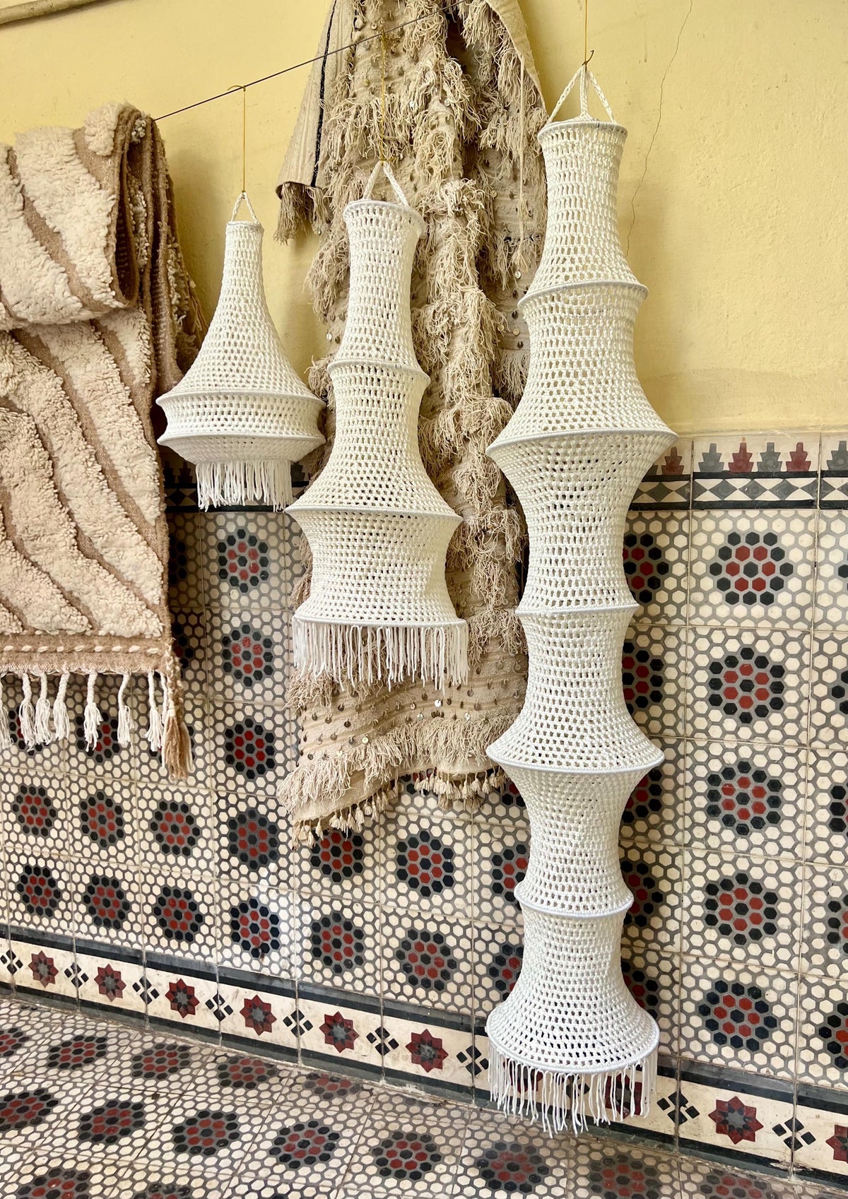 Three off-white crocheted hanging lampshades of varying heights, each featuring a tiered, open-weave structure with a…