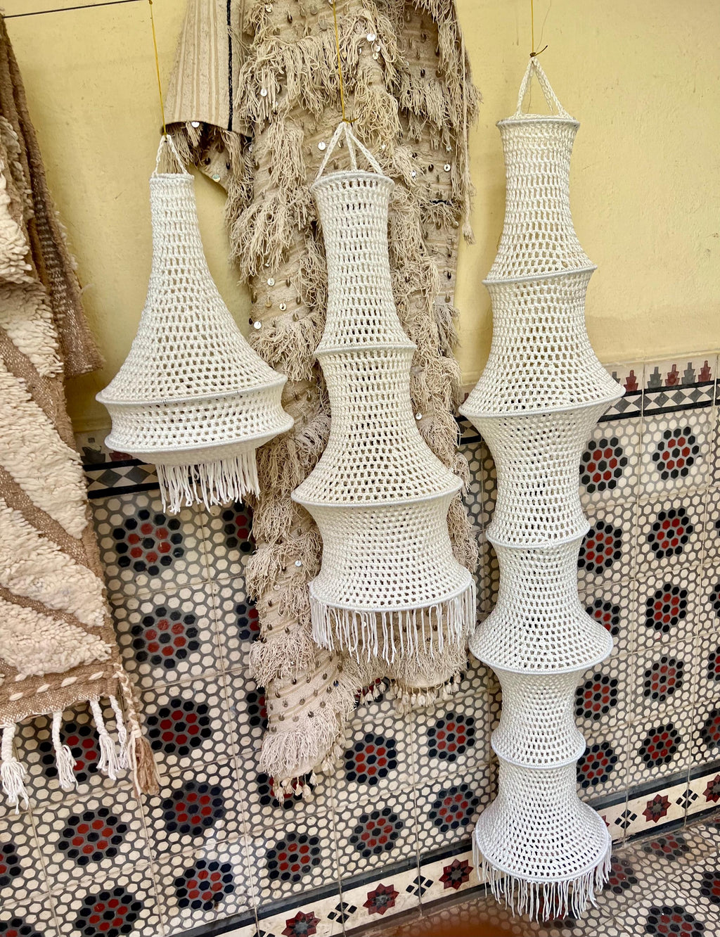 Three white crocheted hanging lampshades of varying heights and tiered shapes. Each lamp features an open-weave pattern…