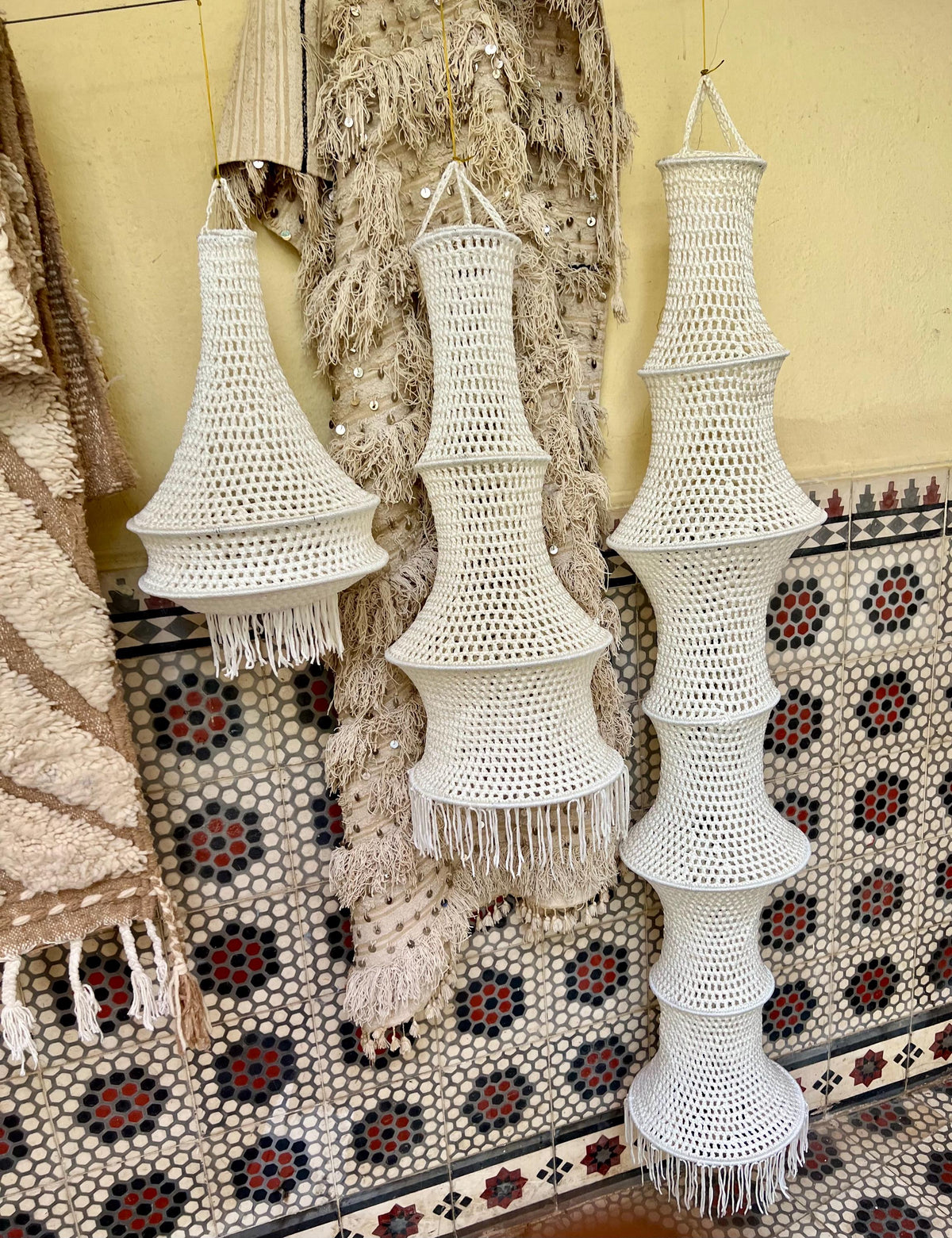 Three white crocheted hanging lampshades of varying heights and tiered shapes. Each lamp features an open-weave pattern…