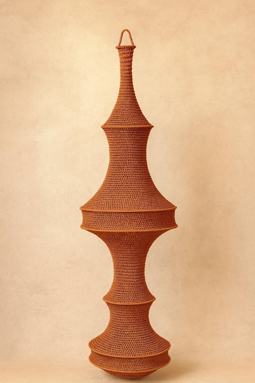 A tall, vertically oriented decorative object crafted from rust-colored yarn in a dense crochet stitch. The structure…