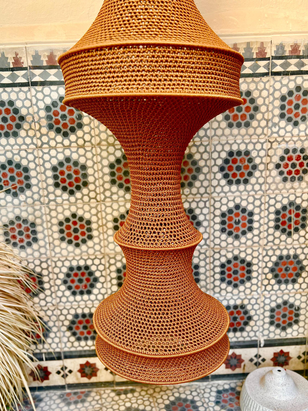 A tall, hourglass-shaped pendant light fixture crafted from a dense, rust-colored woven material. The structure features…