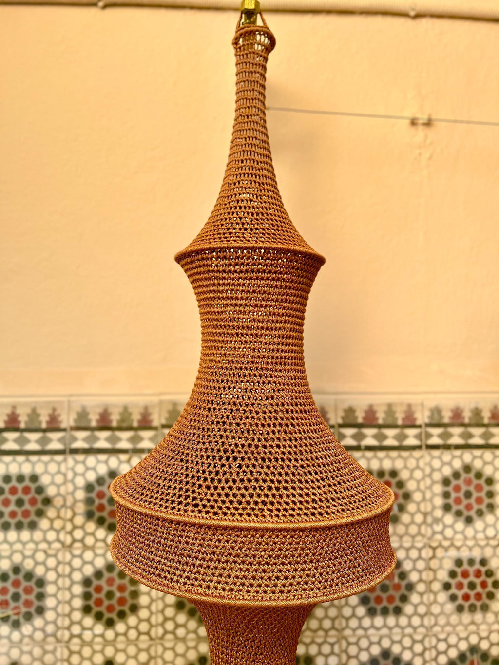 A tall, bell-shaped pendant light fixture featuring a brown, tightly woven crochet or knit texture. The structure tapers…