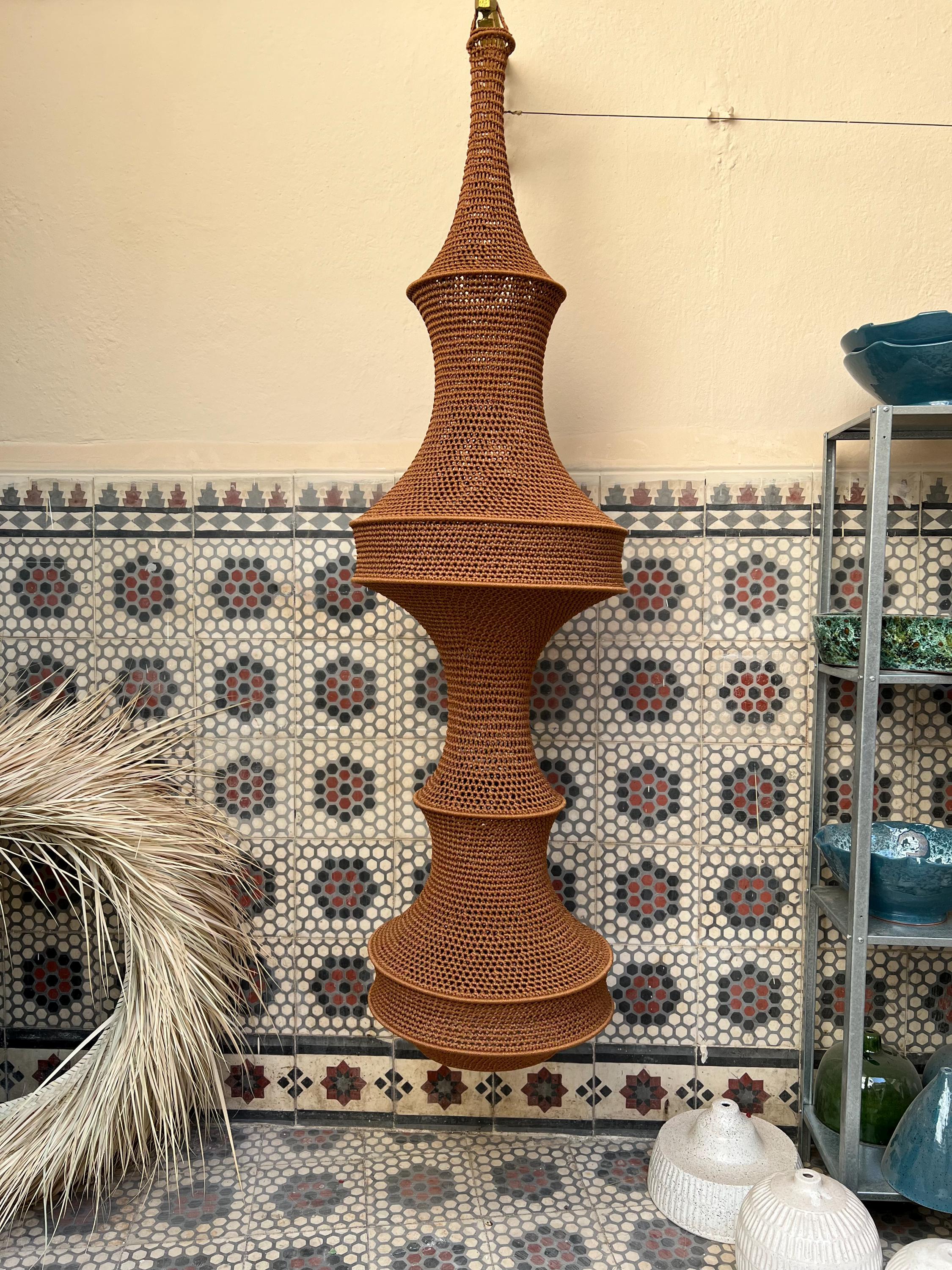 A tall, vertically oriented hanging pendant light fixture featuring a brown, crocheted or woven fiber shade with a…
