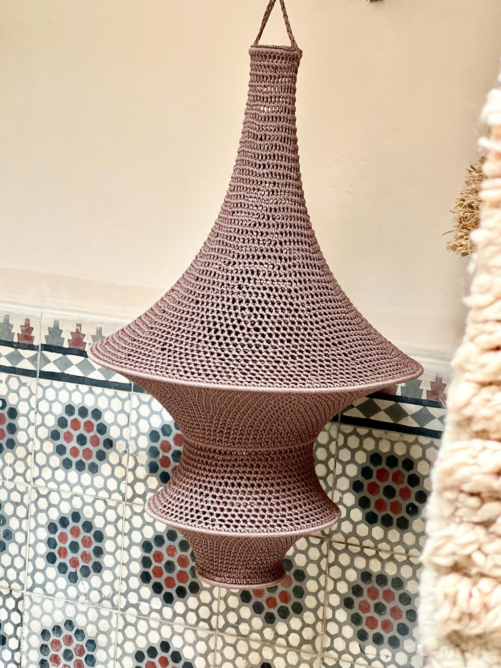 A mauve-colored, hand-crocheted pendant light fixture with a distinct hourglass shape. The structure features a wide,…