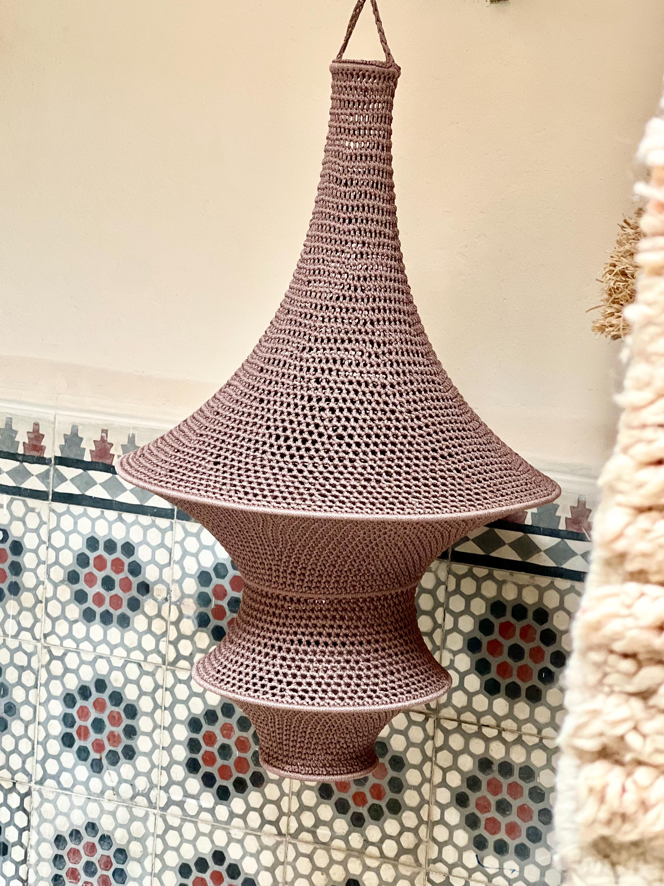A mauve-colored, hand-crocheted pendant light fixture with a distinct hourglass shape. The structure features a wide,…