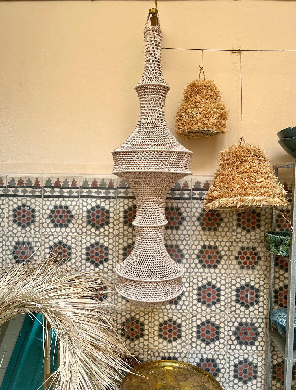 A tall, beige, crocheted pendant light fixture with a multi-tiered, hourglass-inspired silhouette. The structure features…