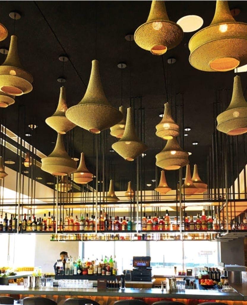 A collection of bell-shaped pendant lights with a woven, light-tan texture suspended from a dark ceiling above a bar…