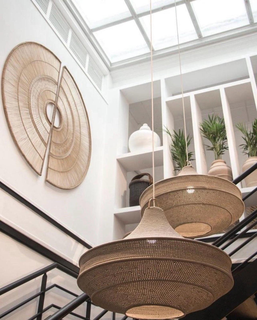 Two large, tiered, woven tan pendant lights hang from the ceiling. On the white wall, a large circular wall art piece is…