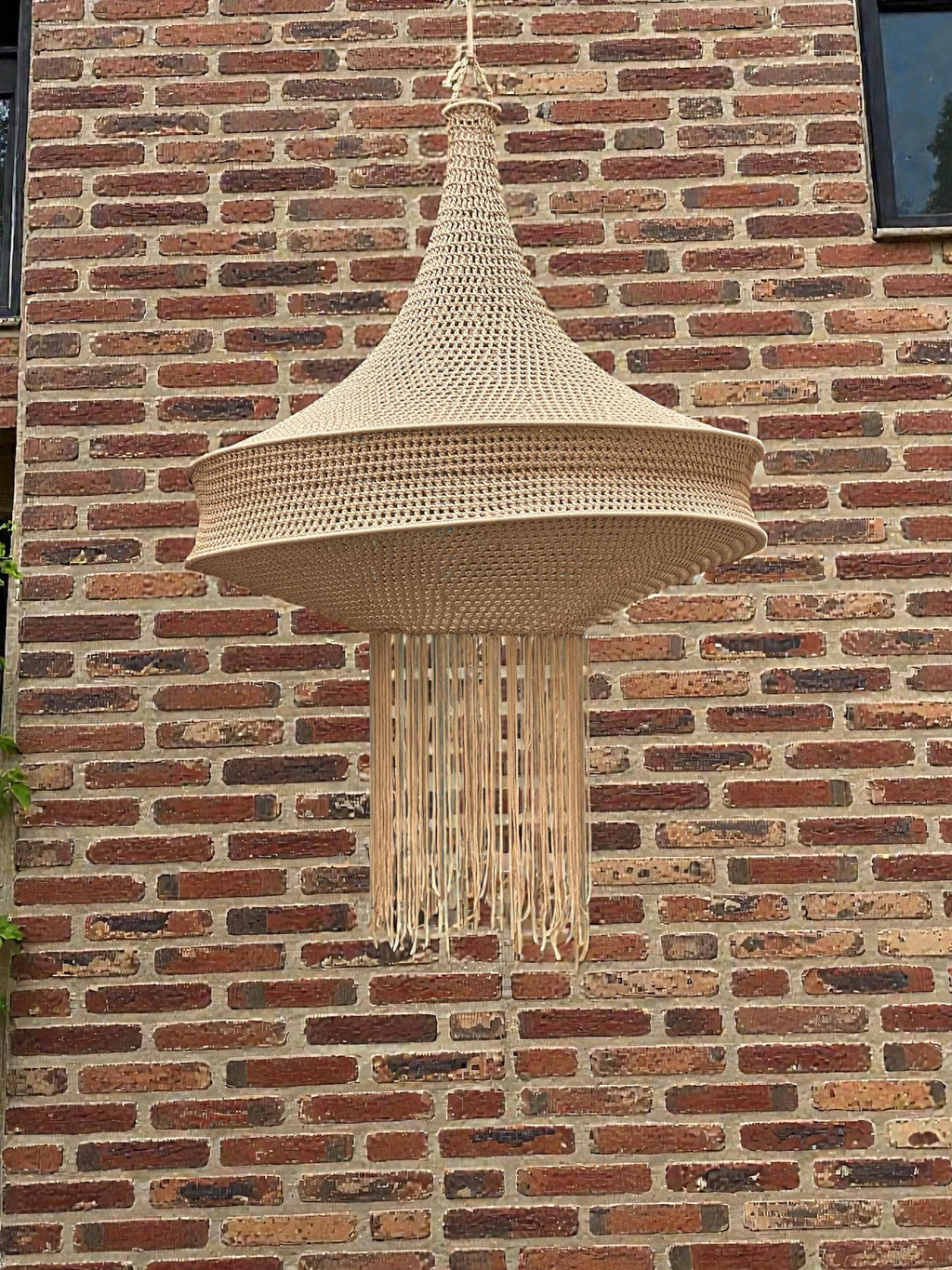 A beige crochet pendant light fixture featuring a wide, tiered conical shade with a textured, woven pattern. The bottom…