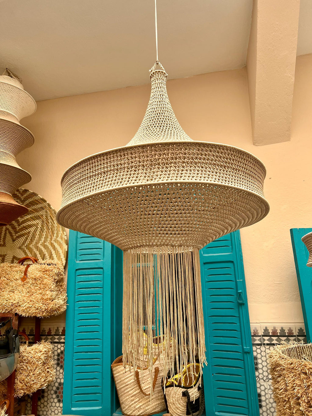 A beige hanging pendant light fixture featuring a wide, circular, crocheted shade with a tapered top and a long, dangling…