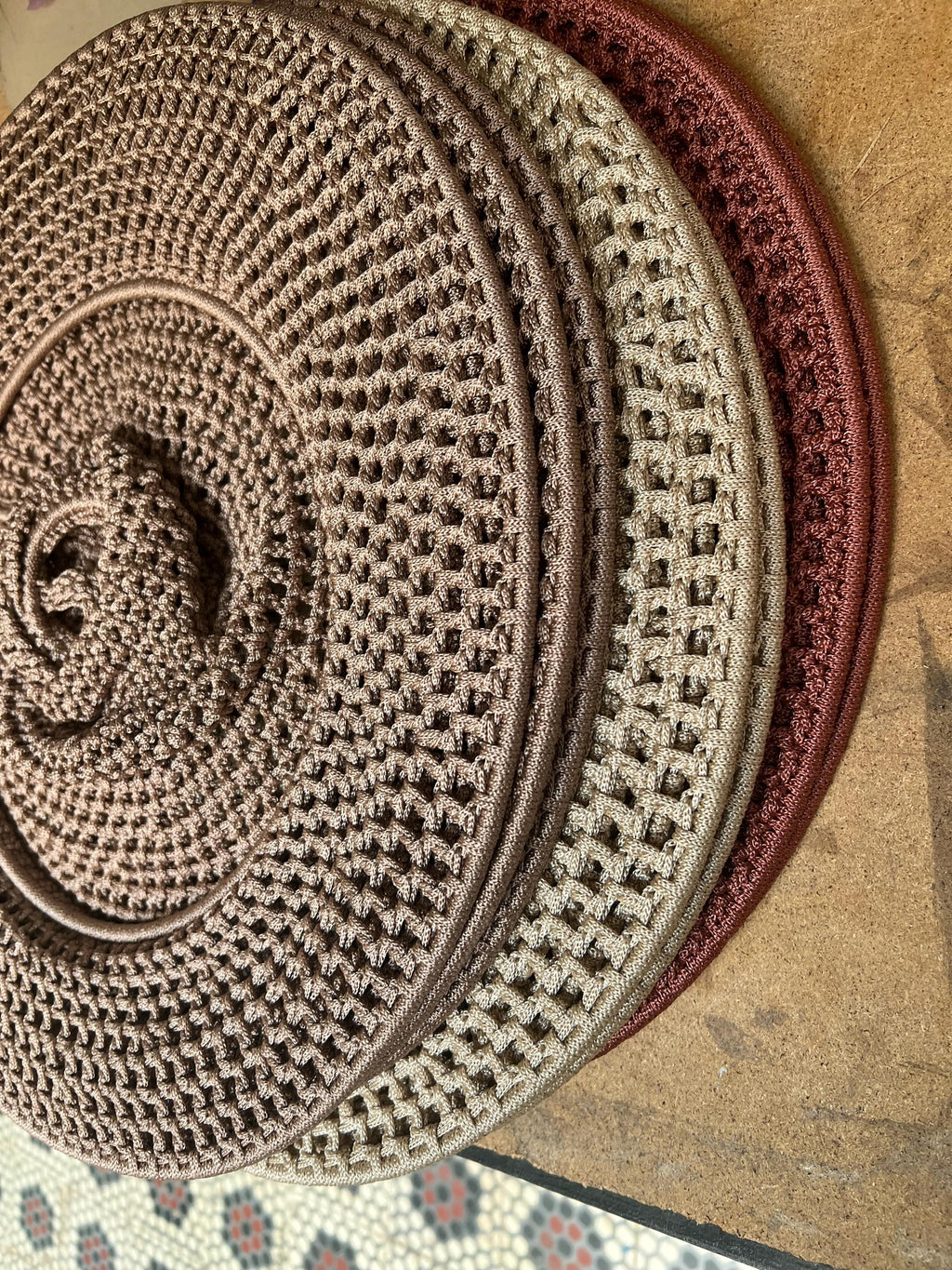 A stack of four circular, crocheted coasters in varying shades of brown, tan, and copper. Each coaster features a dense,…