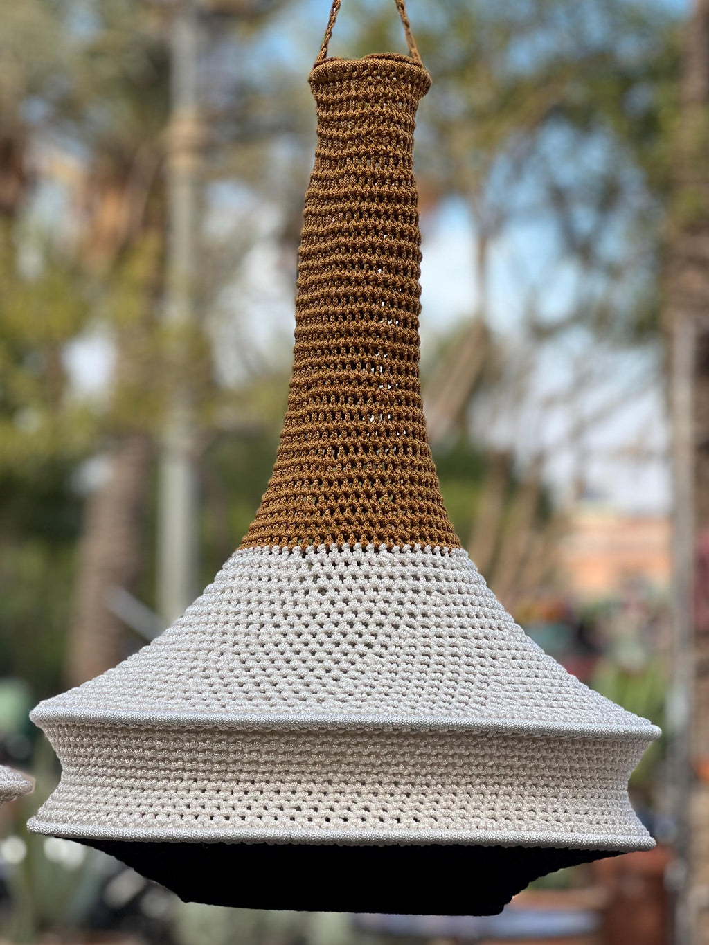 A hanging pendant light shade featuring a tall, narrow neck crocheted in a warm brown yarn that transitions into a wide,…