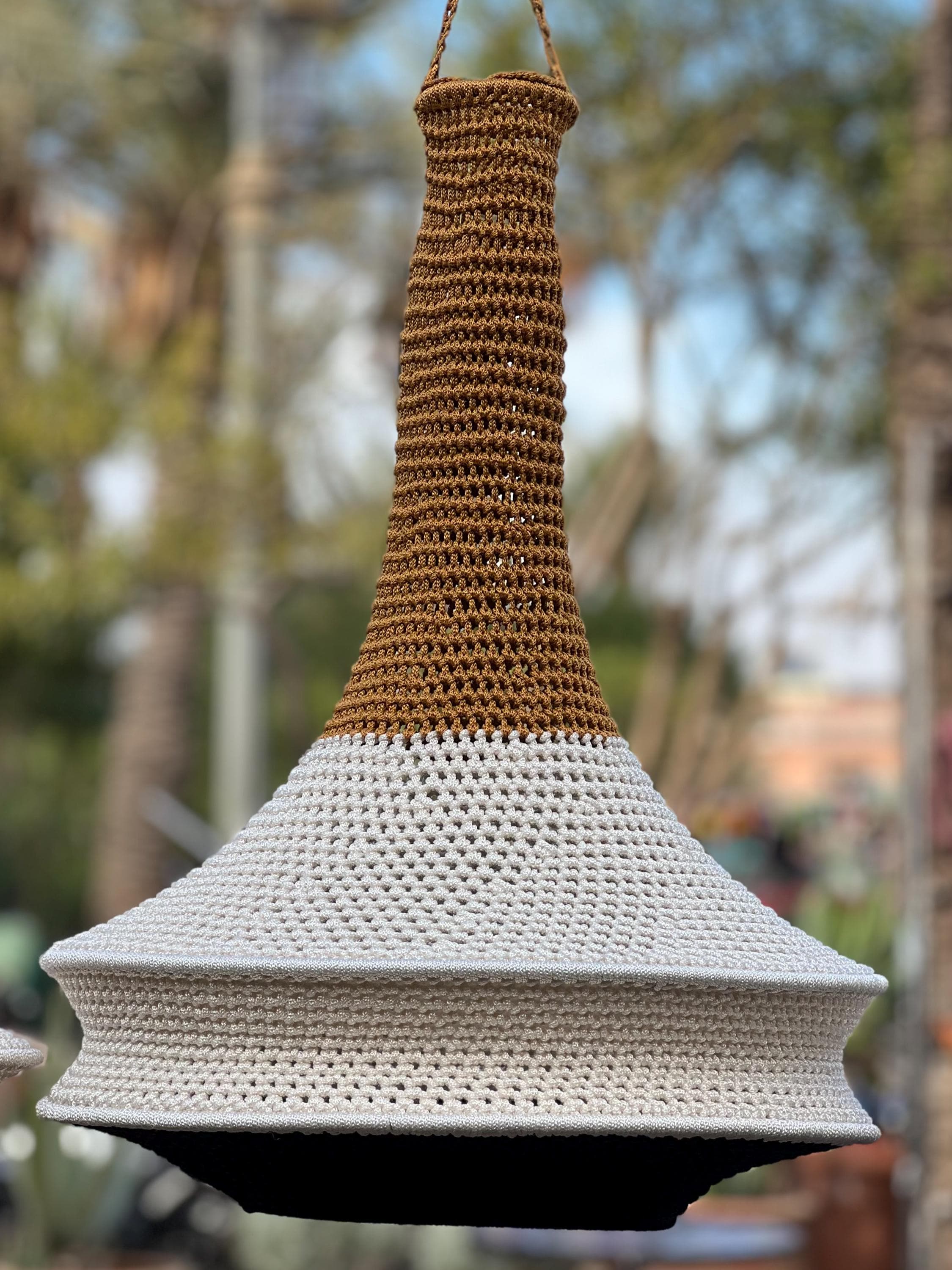 A hanging pendant light shade featuring a tall, narrow neck crocheted in a warm brown yarn that transitions into a wide,…