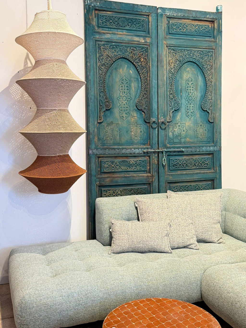 A multi-tiered pendant light in shades of beige and brown hangs in front of a teal carved wooden door. A light blue…