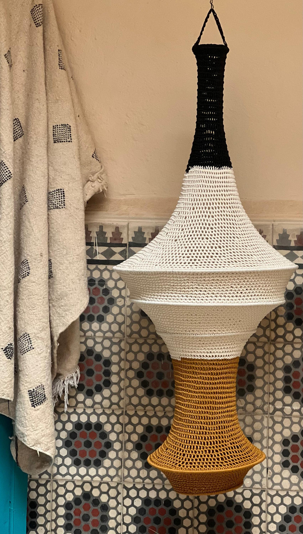 A hanging pendant light shade featuring a crocheted design with a black top section, a wide white middle section, and a…