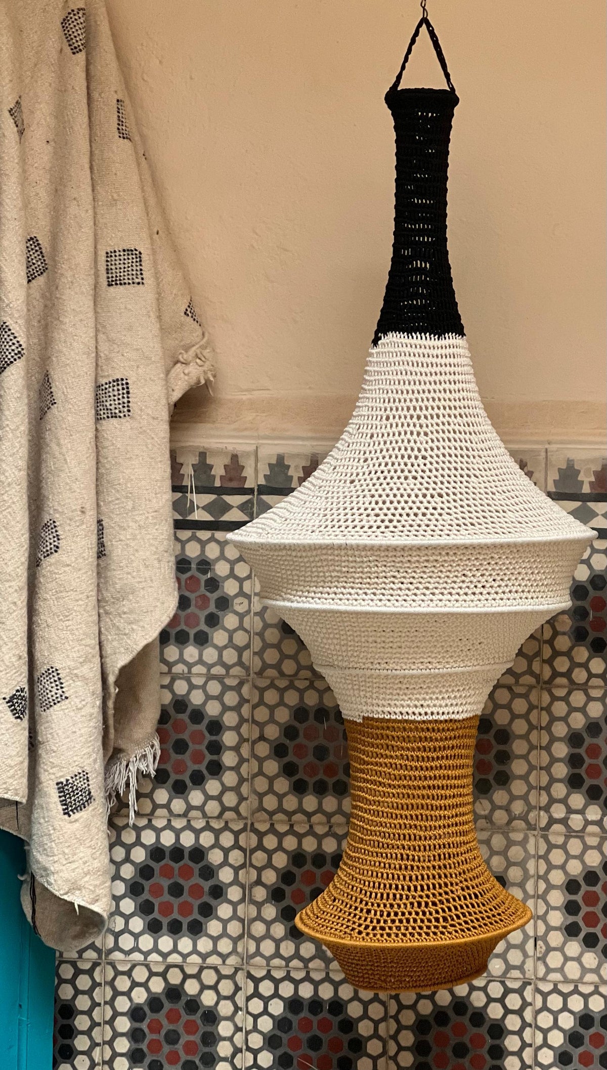 A hanging pendant light shade featuring a crocheted design with a black top section, a wide white middle section, and a…