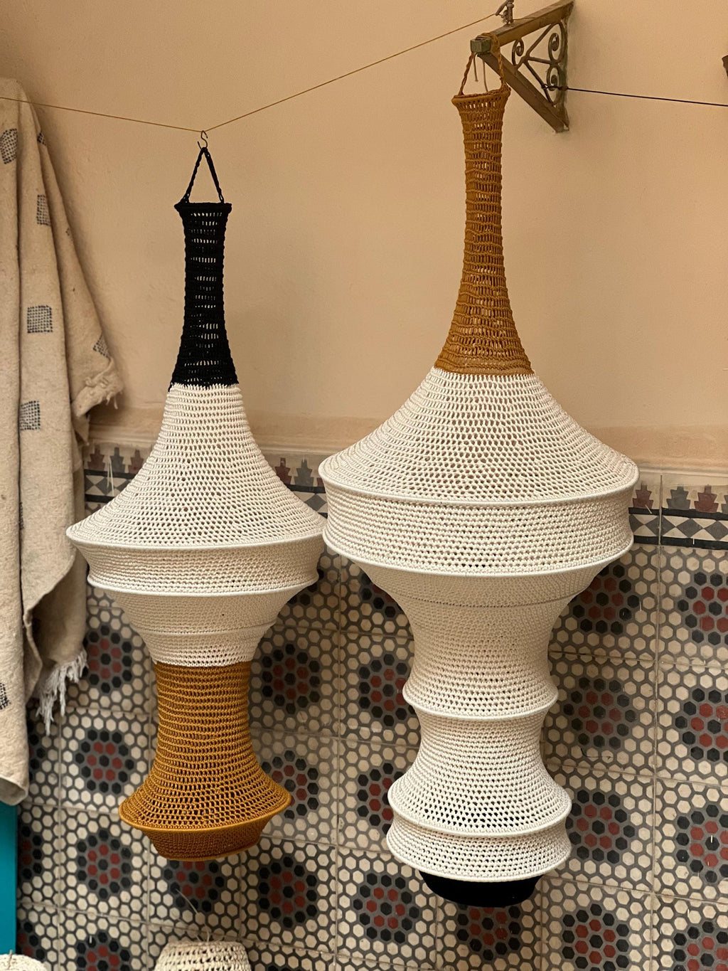 Two hanging pendant lamps crafted from crocheted fiber. The left lamp features a black top, white middle, and mustard…
