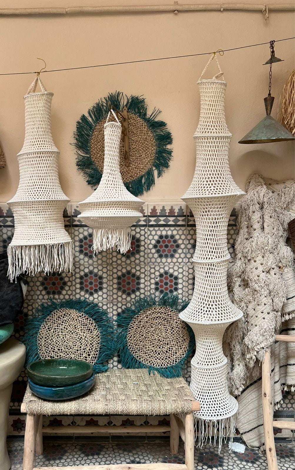 Three off-white crocheted hanging lampshades with open-weave patterns and fringe, two round wall hangings with teal…
