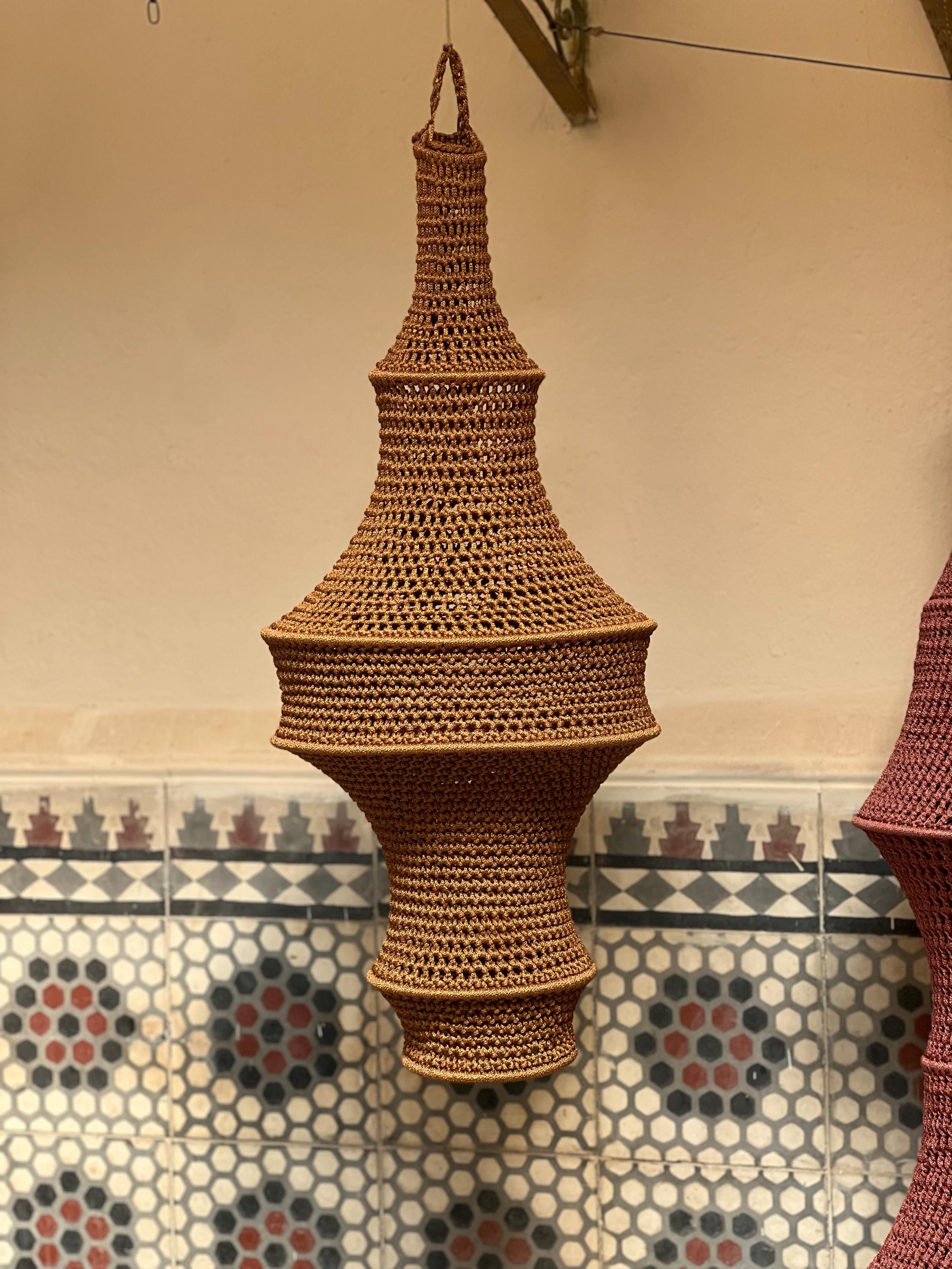 A hanging light fixture shade crafted from brown crocheted yarn. The shade features a multi-tiered, bulbous silhouette…