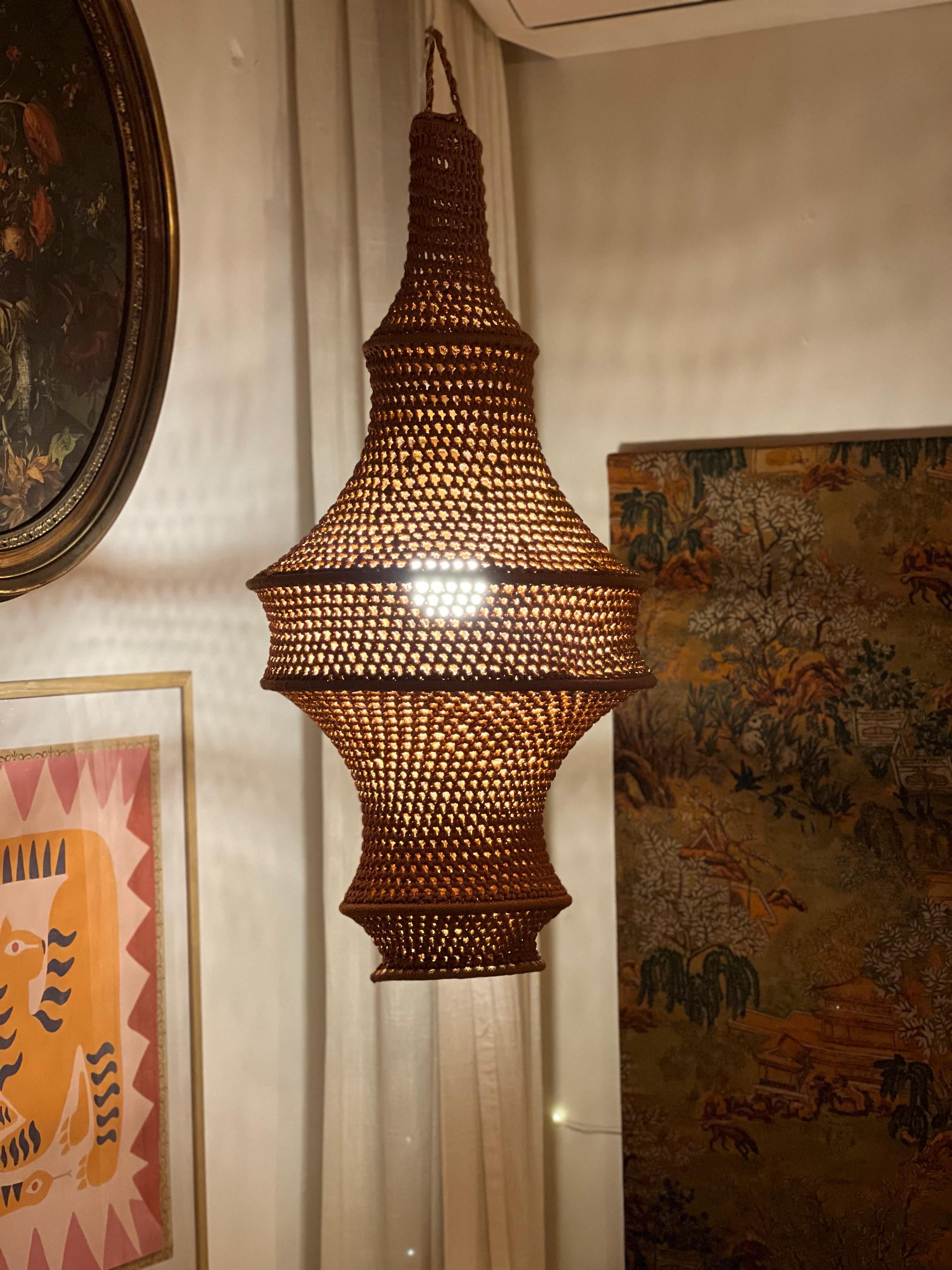 A brown, woven, bell-shaped pendant light fixture with a complex, open-work lattice pattern. The structure features a…