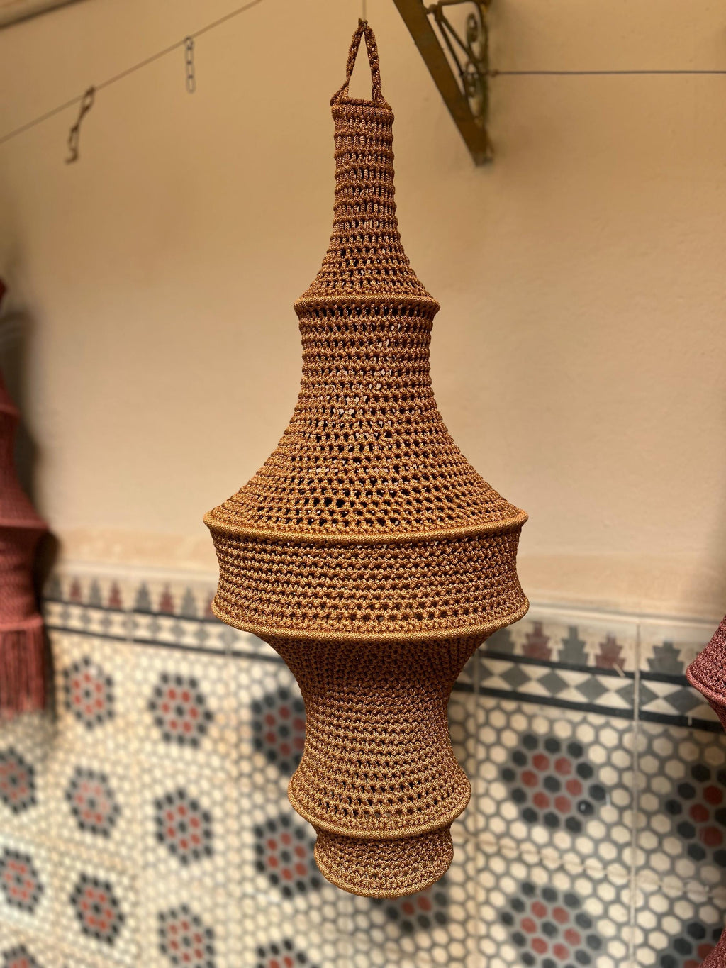 A hanging light fixture shade crafted from brown yarn in a crochet technique. The shade features a multi-tiered,…