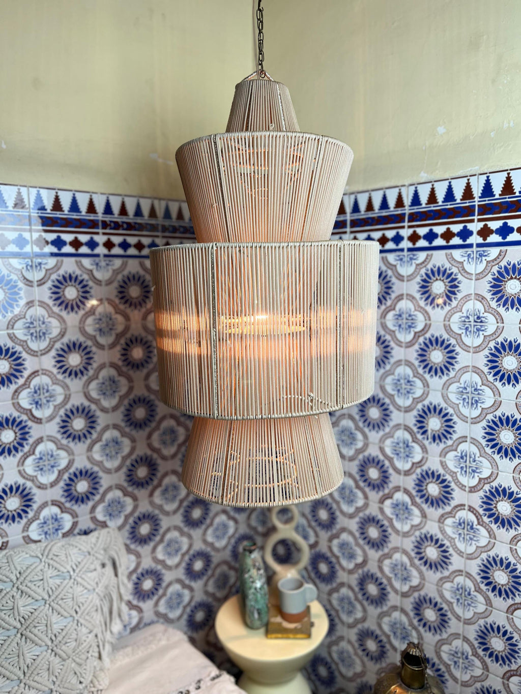 A beige, tiered pendant light fixture. The light is made of vertical, thin, light-colored rods, creating a layered, open…