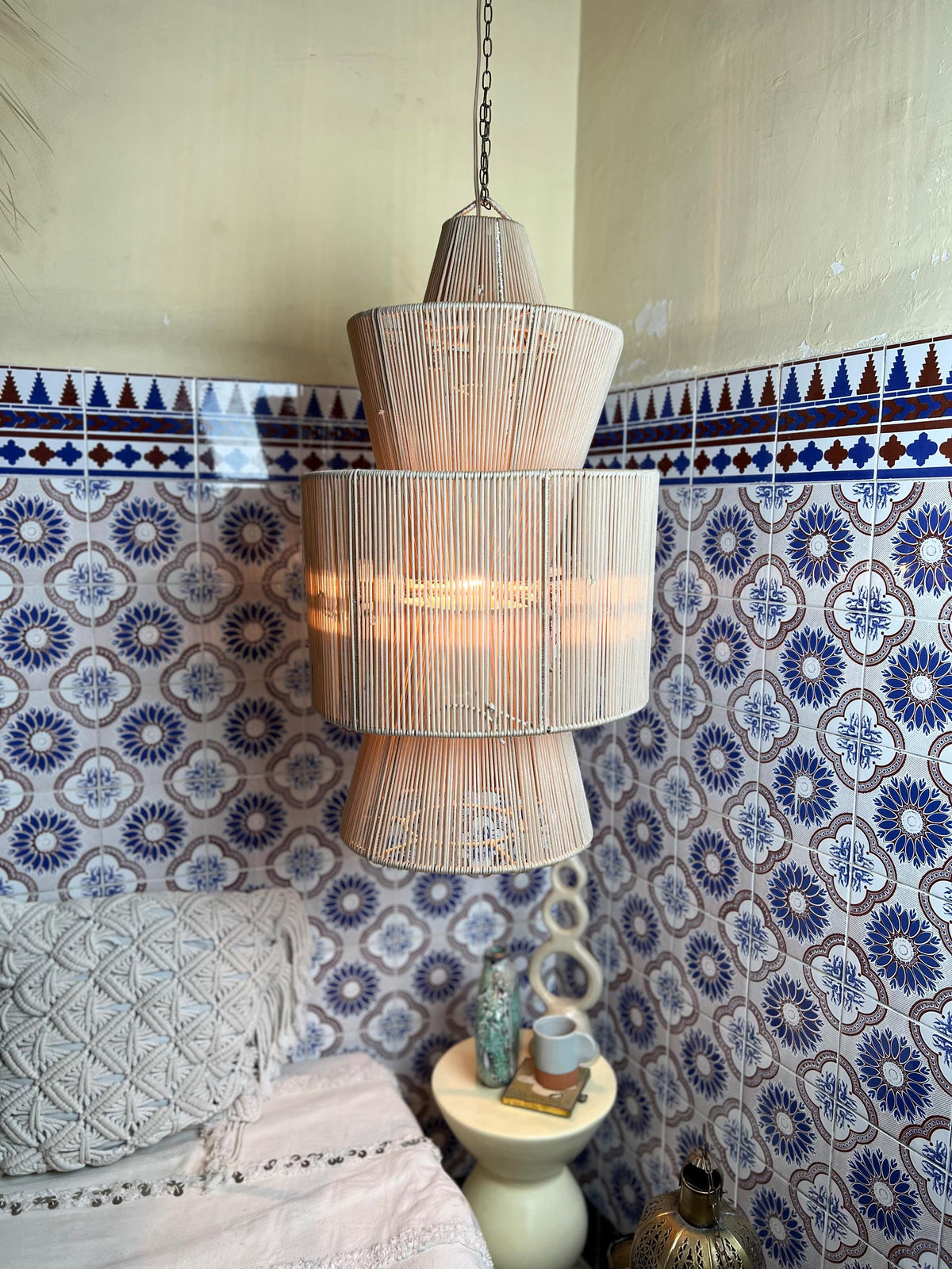 A three-tiered, cream-colored pendant light fixture. The light is suspended from a chain and features a woven design. The…