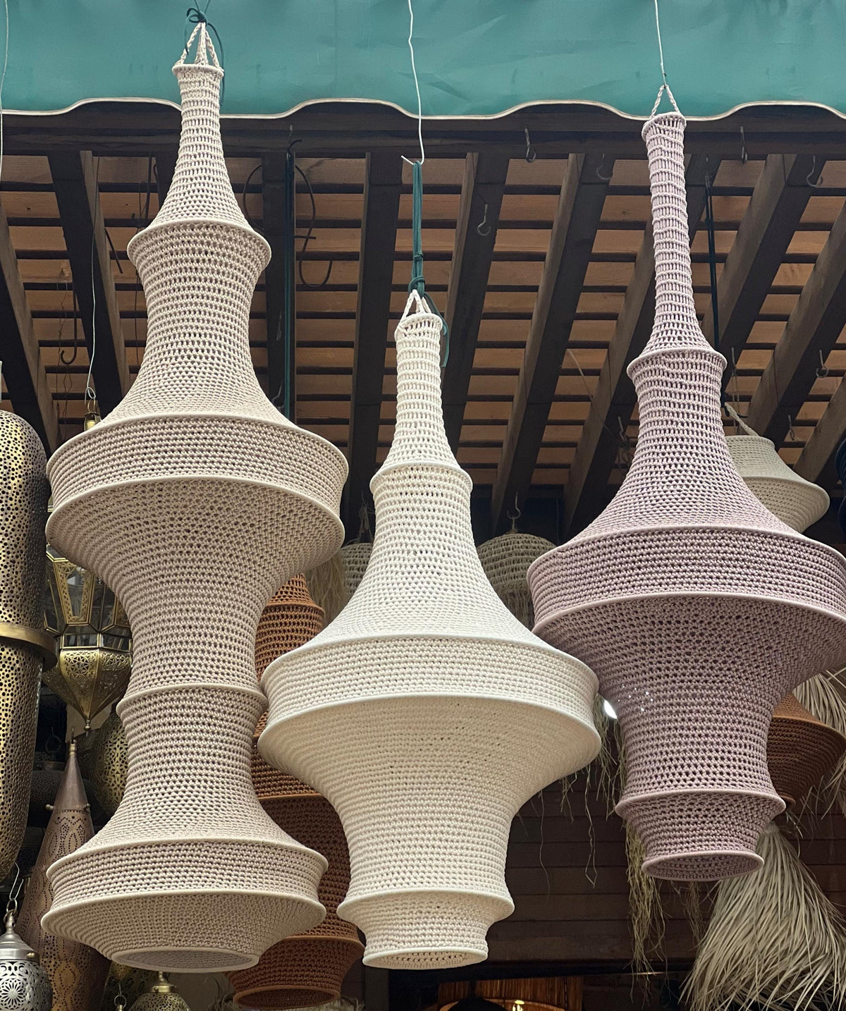 Three hanging pendant lamps featuring intricate crochet work in soft beige, off-white, and light mauve. Each lamp has a…