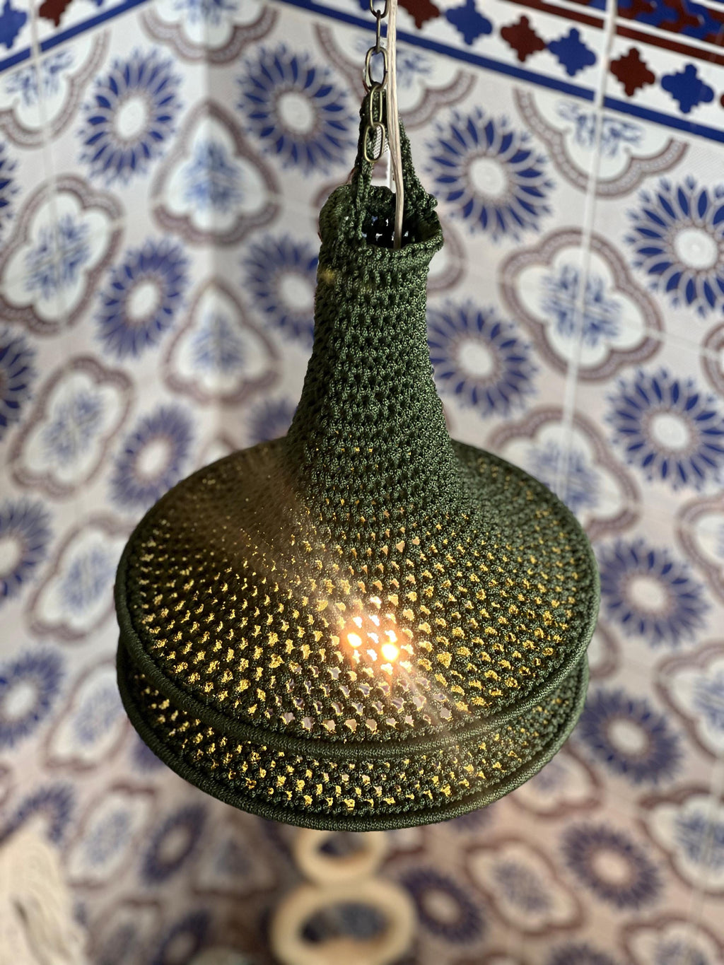 A dark olive green crocheted pendant light fixture with a bell-shaped shade. The textured fabric features a dense,…