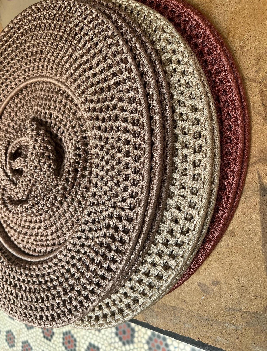 A stack of three circular, crocheted coasters or mats. The top piece is a dark brown, the middle is a light beige, and…