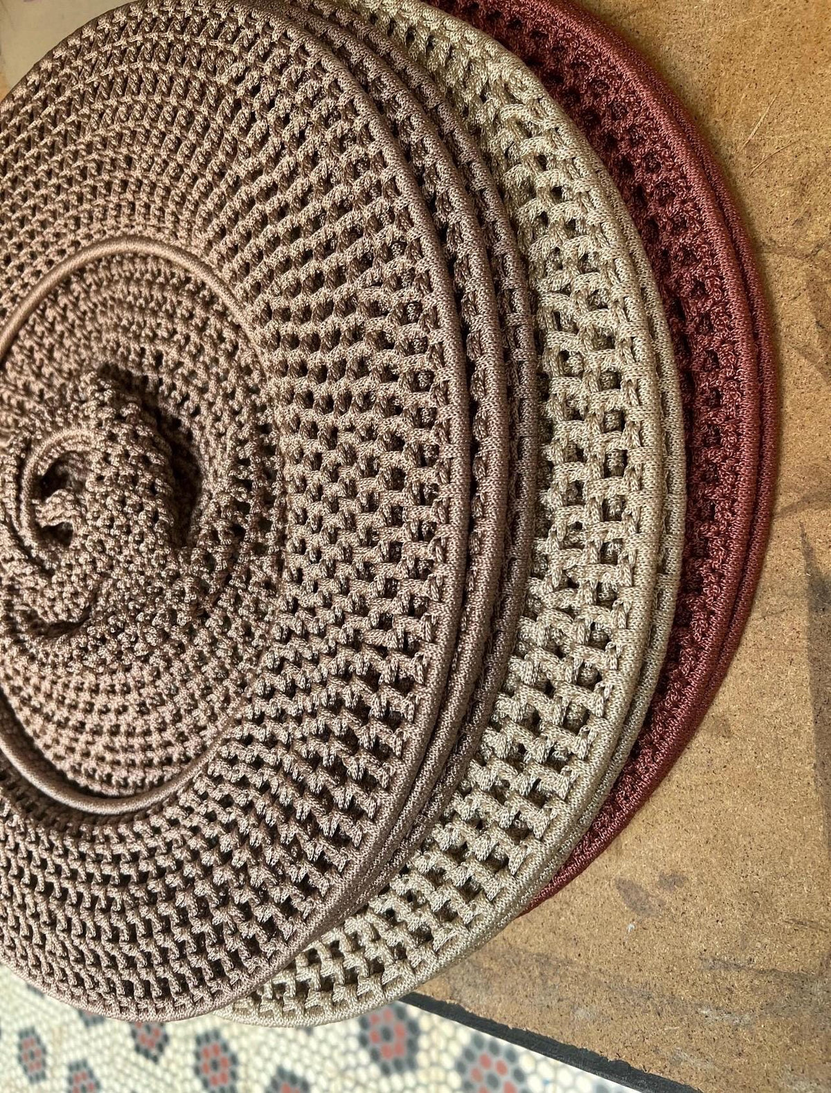 A stack of three circular, crocheted coasters or mats. The top piece is a dark brown, the middle is a light beige, and…