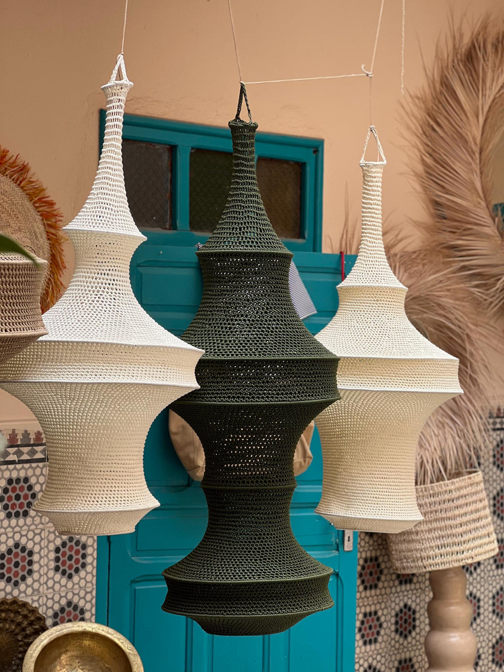 Three hanging pendant lamps featuring intricate crochet work. Two lamps are off-white, and one central lamp is dark olive…