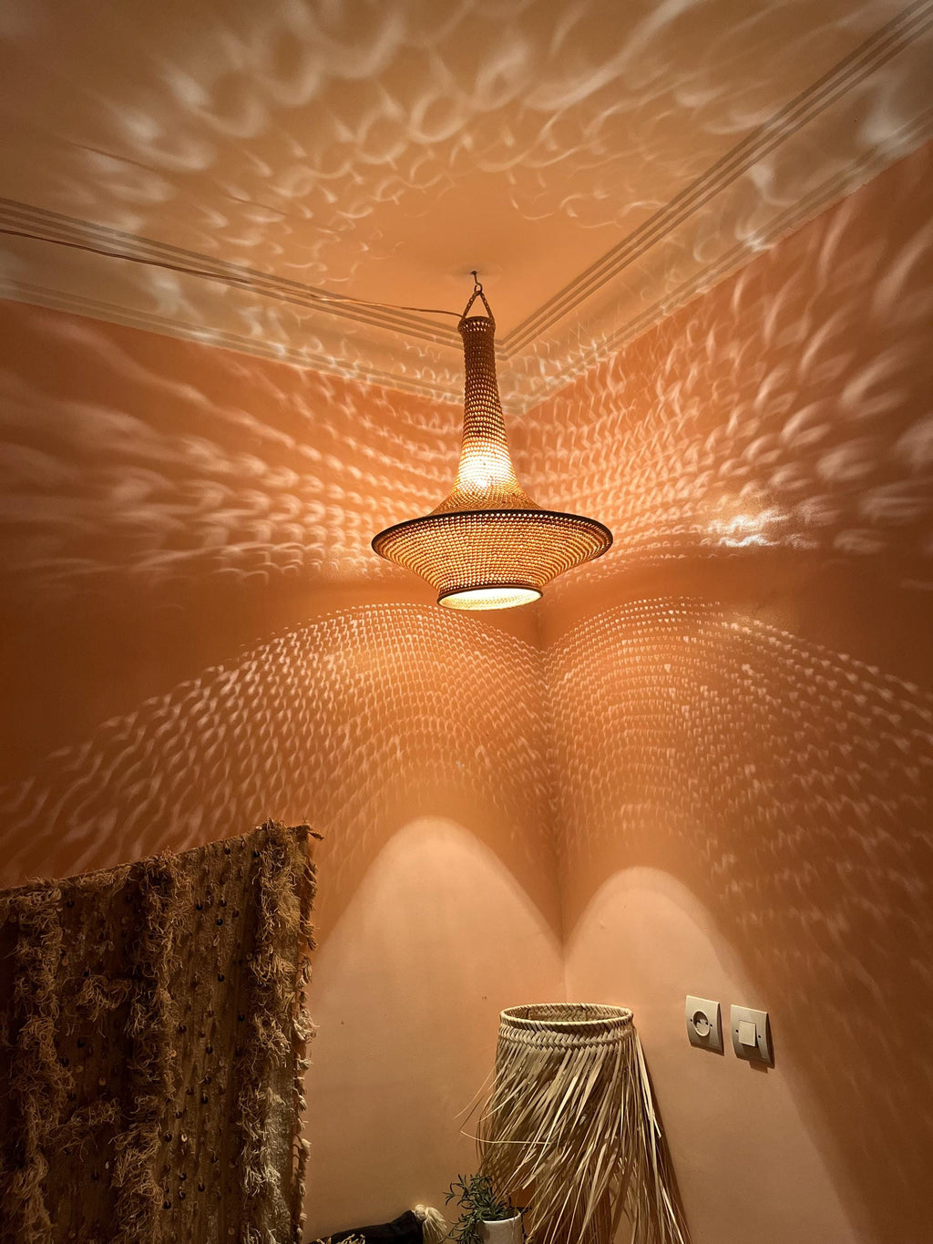 A woven pendant light fixture with a conical top and a wide, circular base hangs from the ceiling. The light casts a…