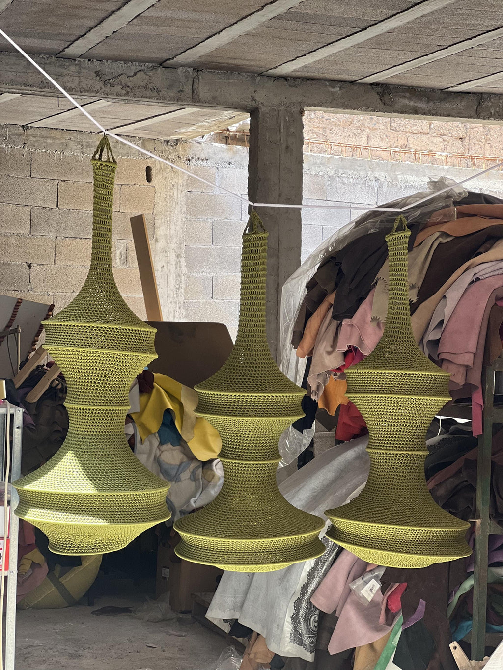 Three olive green, crocheted, bell-shaped pendant light shades hanging from a white cord. Each shade features a narrow,…